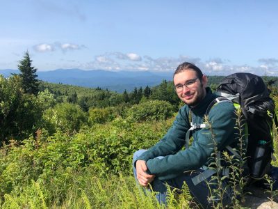 Person in a hiking pack sitting in green grass with a blue mountain in the background.