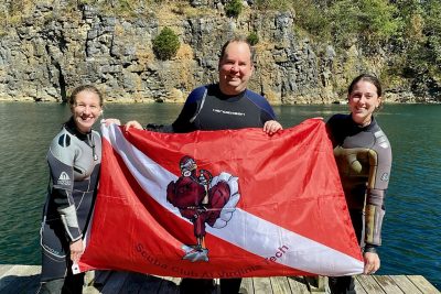 Three people in scuba gear holding a Virginia Tech Hokies Flag while standing in front of a quarry.