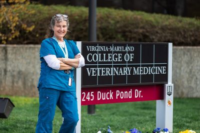 A lady in scrubs standing outside the teaching hospital leaning againgst the sign.